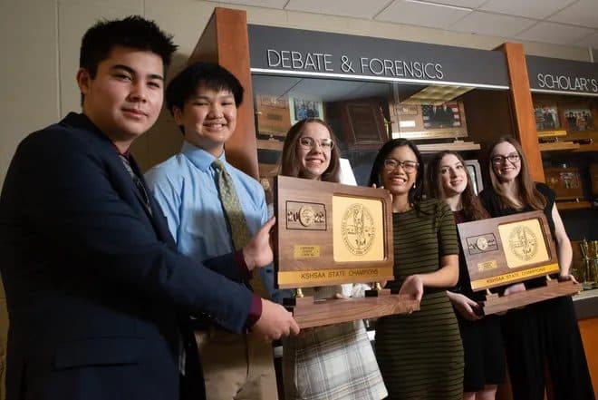 Students holding award plaques in front of a Debate and Forensics trophy case