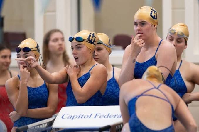 A girls swim team gathers at the pool’s edge mid-meet